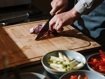 A chef skillfully slices onions on a wooden board with fresh vegetables ready in bowls.